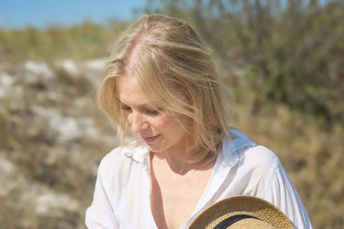 Woman with fine light-blonde shoulder-length hair outdoors in bright summer sun, visible part line on the scalp, straw hat held in one hand