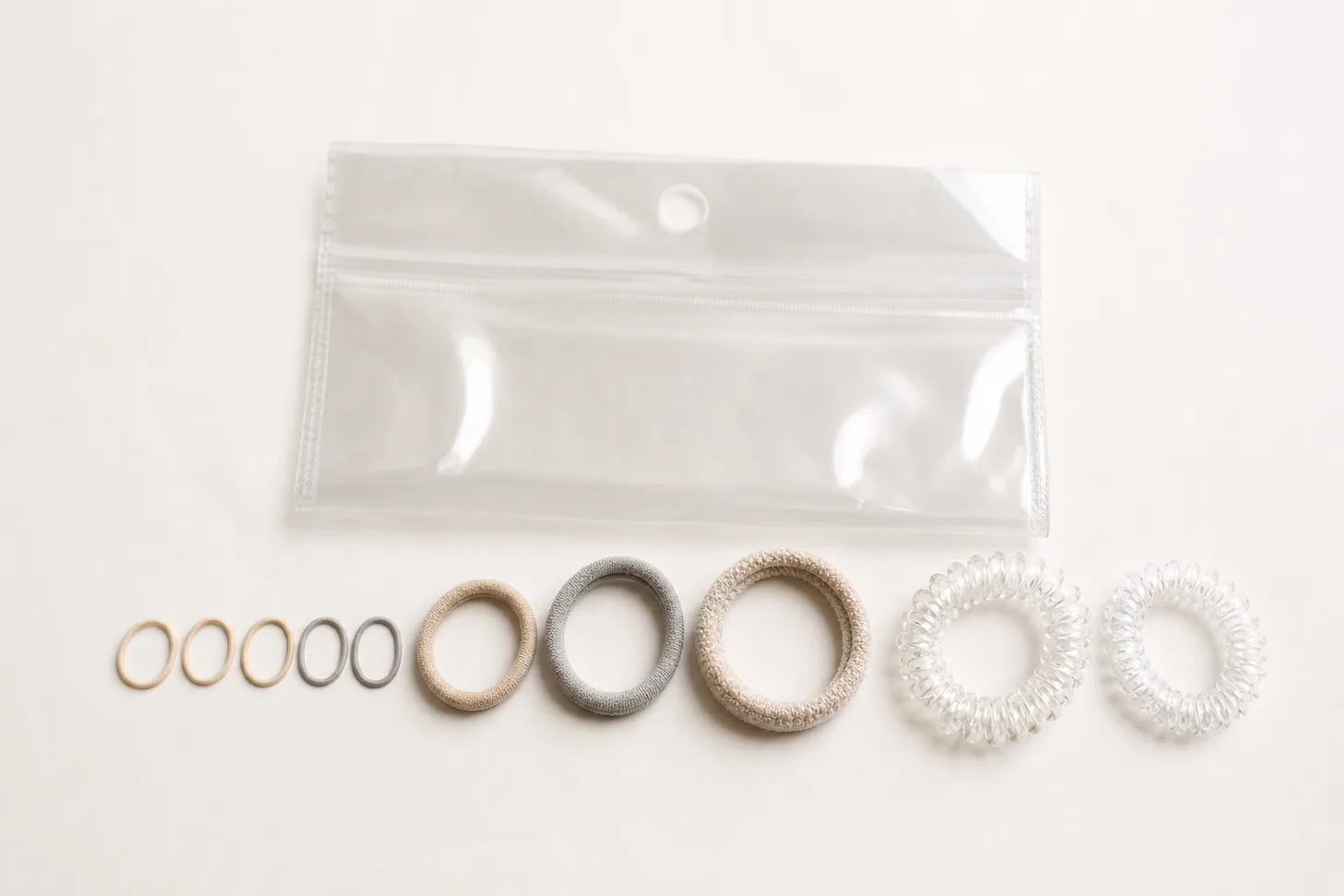 An overhead row of small hair ties, fabric elastics and clear spiral coils on a white surface