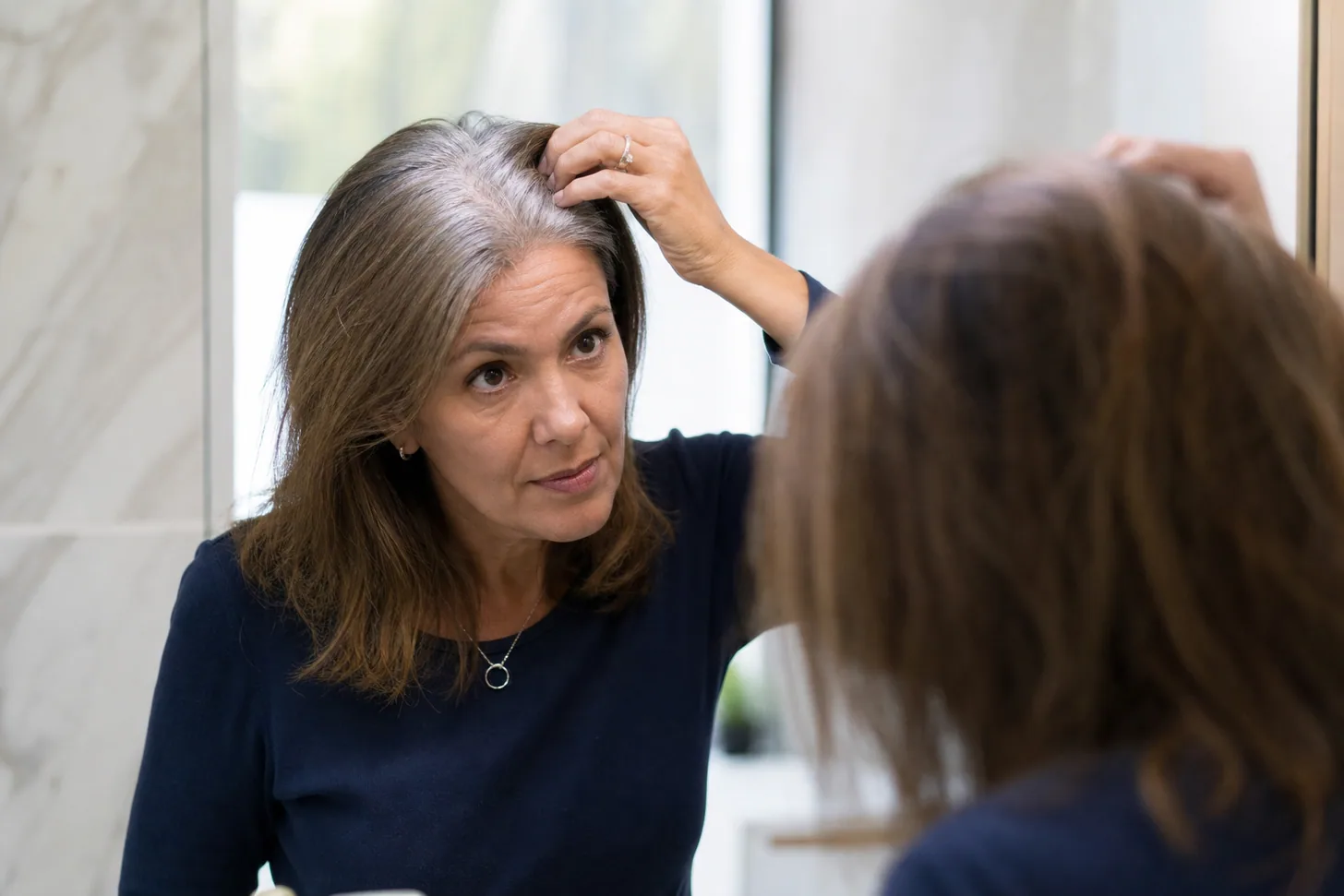 Woman in her early fifties examining her parting in a bathroom mirror in natural daylight, fine mid-length hair with visible gray at the roots, calm and unposed